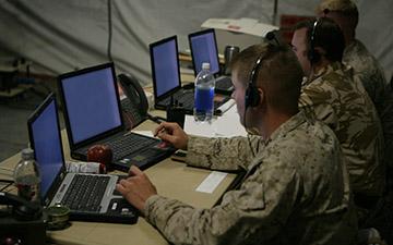 Three male warfighters utilizing satcom to communicate over their headsets, looking at laptops in a tactical operations center